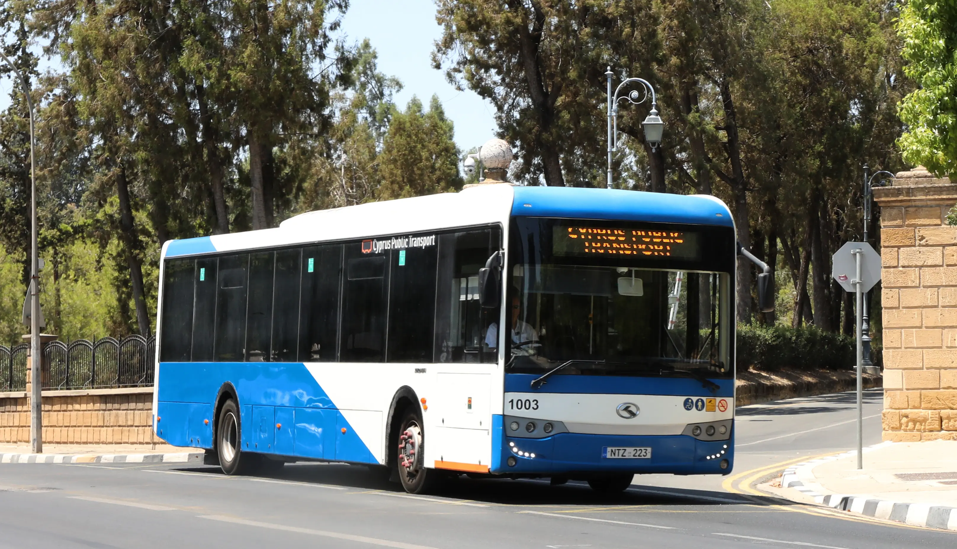 Blue and white Cyprus Public Transport bus driving on a tree-lined street near a stone building and sidewalk.